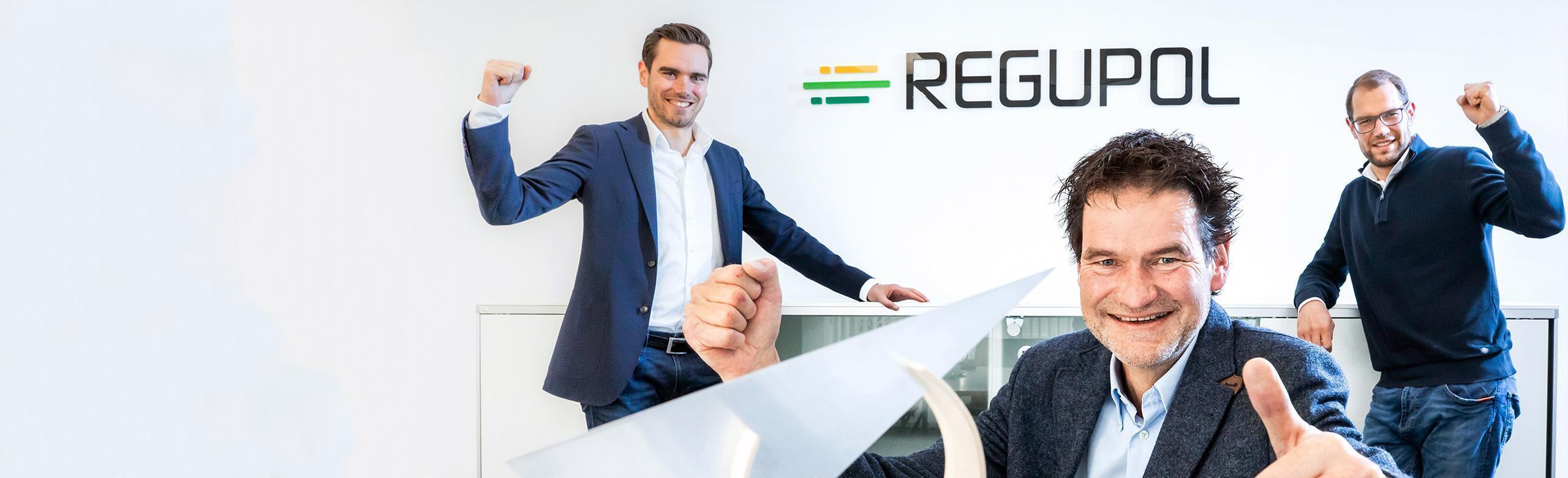 Three men, the CEOs of REGUPOL, stand smiling with raised arms in a bright room in front of the company logo. In the foreground is the Environmental Business Award – a modern, sculptural trophy symbolizing sustainability and achievement.