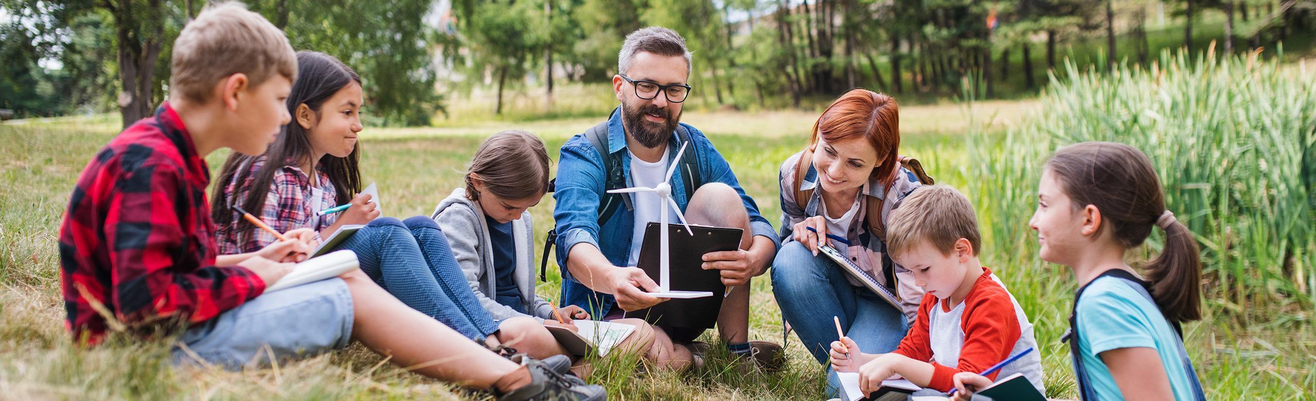 A group of children sits with two adults in a circle on a meadow. They are learning outdoors, using notebooks and a wind turbine model. Trees and reeds can be seen in the background.
