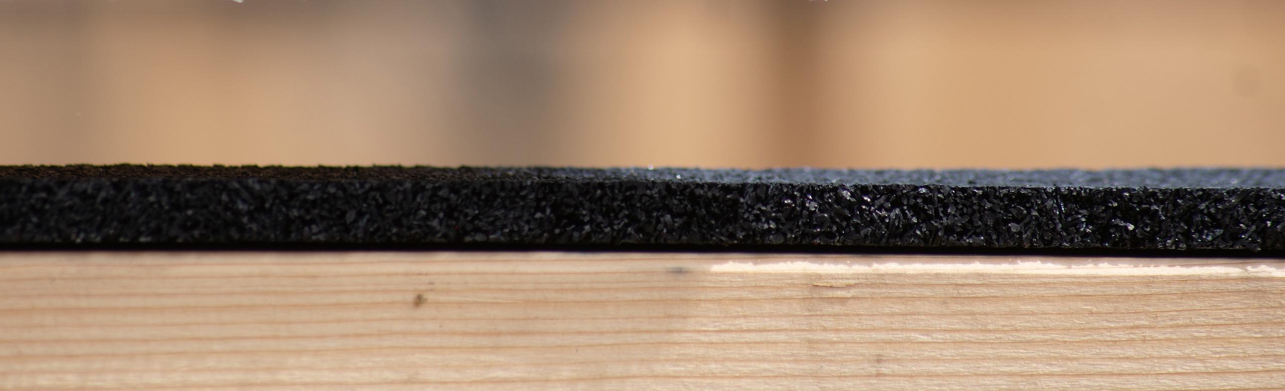 Close-up of a black rubber granulate mat on light wood, shown from the side in cross-section.